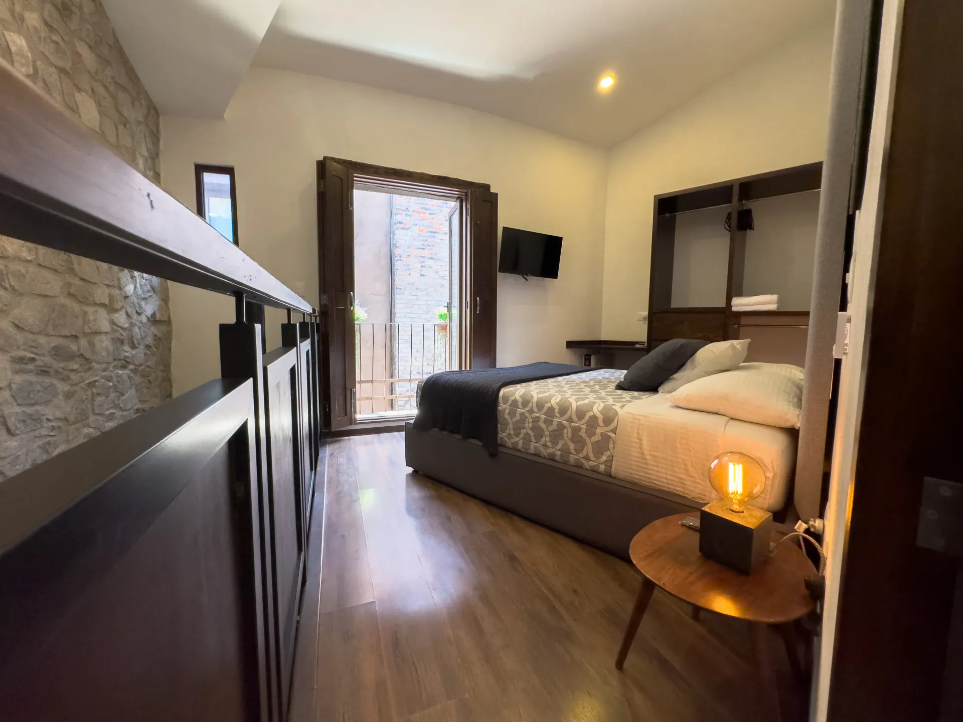 Dormitorio con cama, pared de piedra, detalles de madera y puerta abierta a un patio.