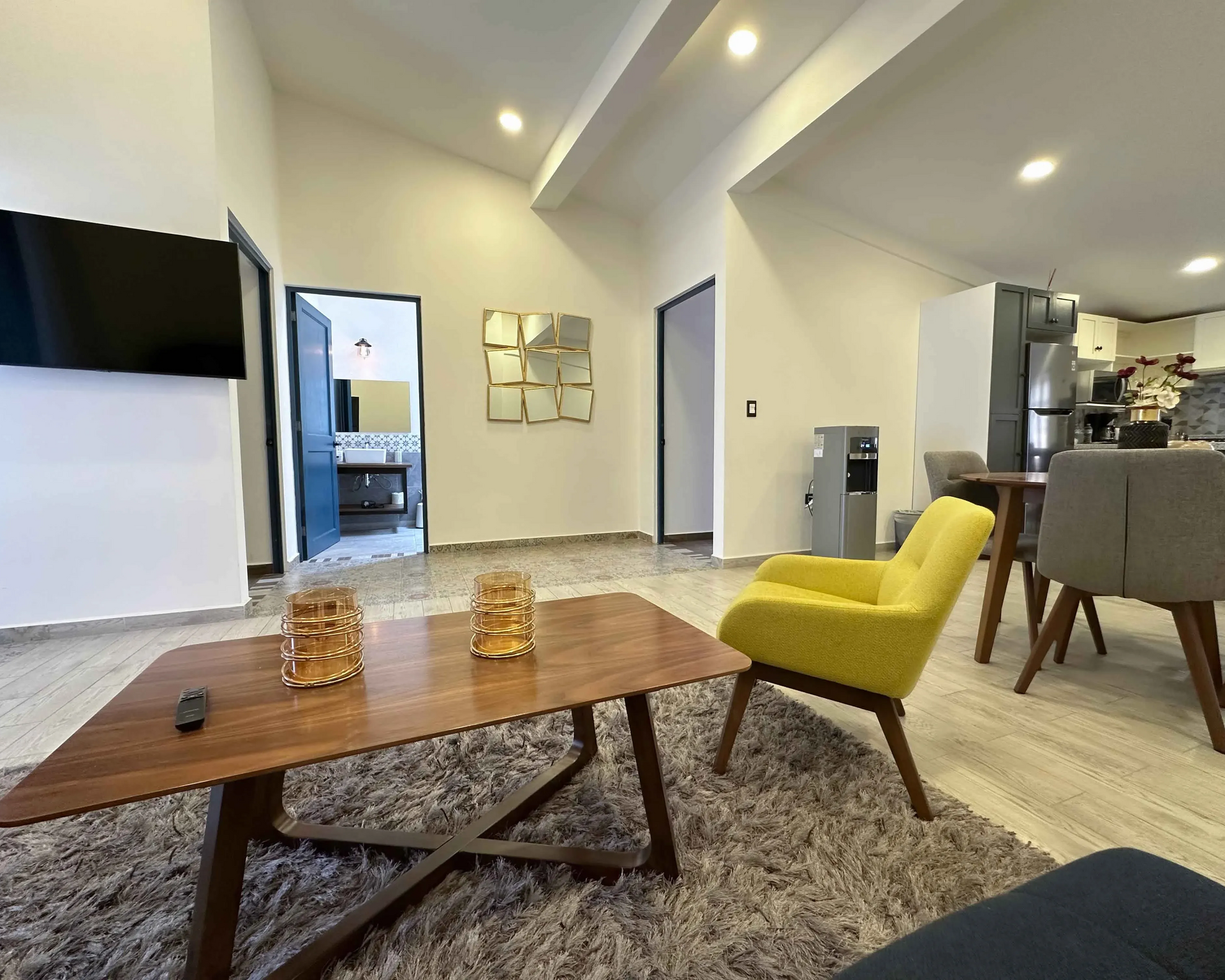 Sala de estar con sillón amarillo, mesa de madera, TV y puerta abierta al baño.