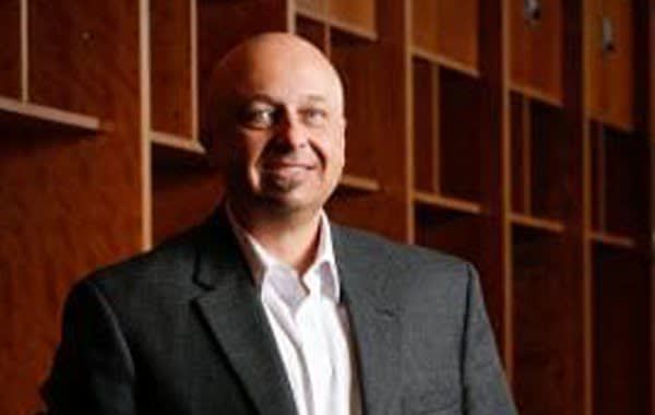 A bald man in a suit stands in front of a wall of wooden shelves