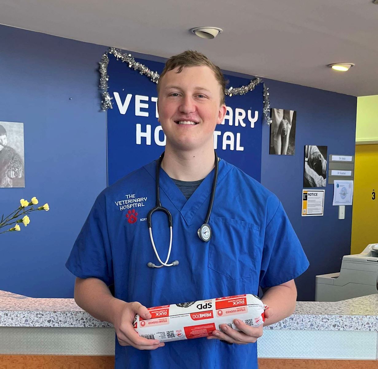 A man in a blue scrub is holding a package in front of a veterinary hospital sign