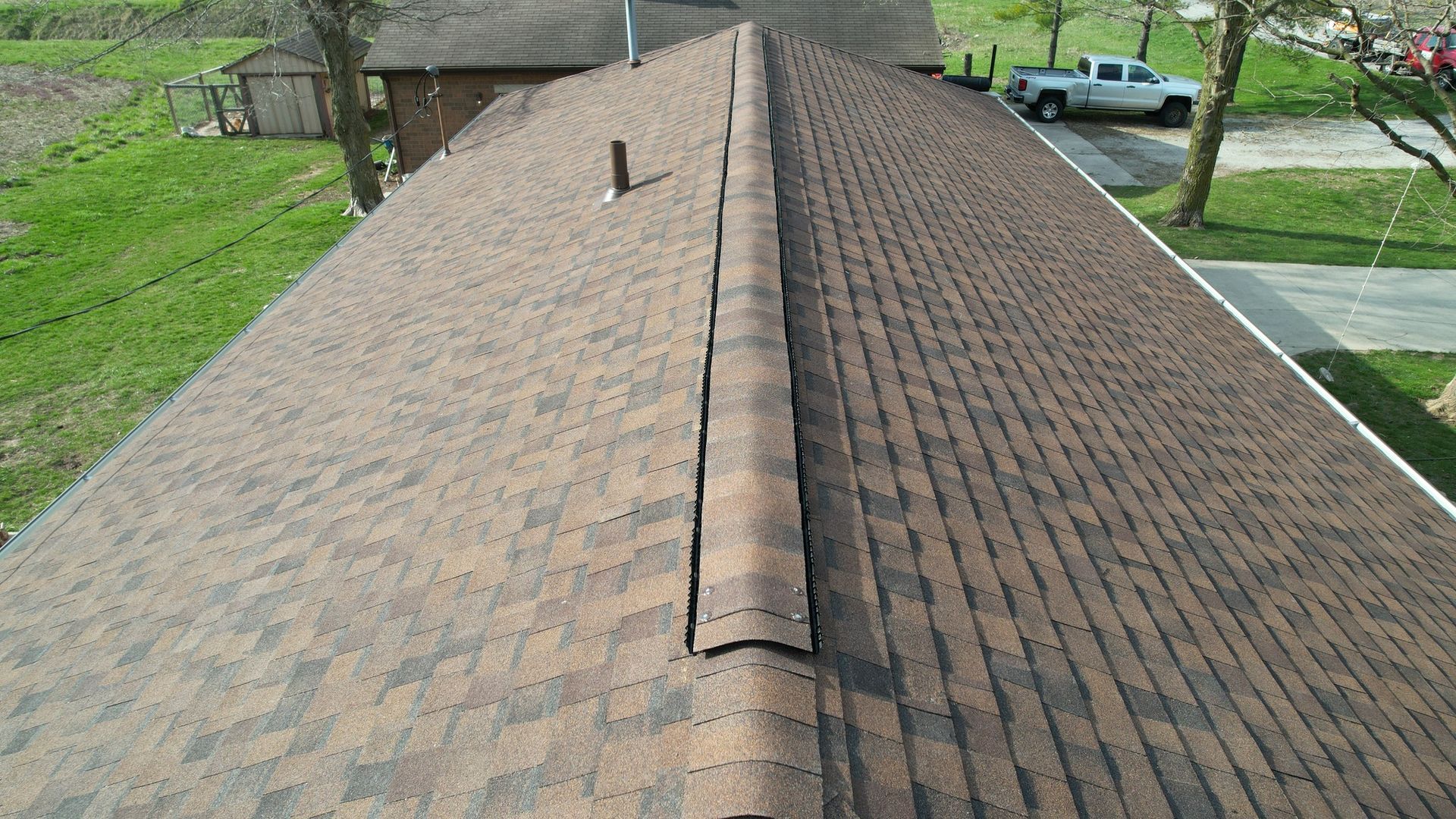 A high-angle view of a brown shingled roof with a central ridge cap, a vent pipe, and a rural background with a truck.