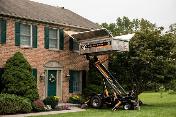A dumpster trailer with hydraulic arms raised, positioning a metal container to roof level at a brick house.