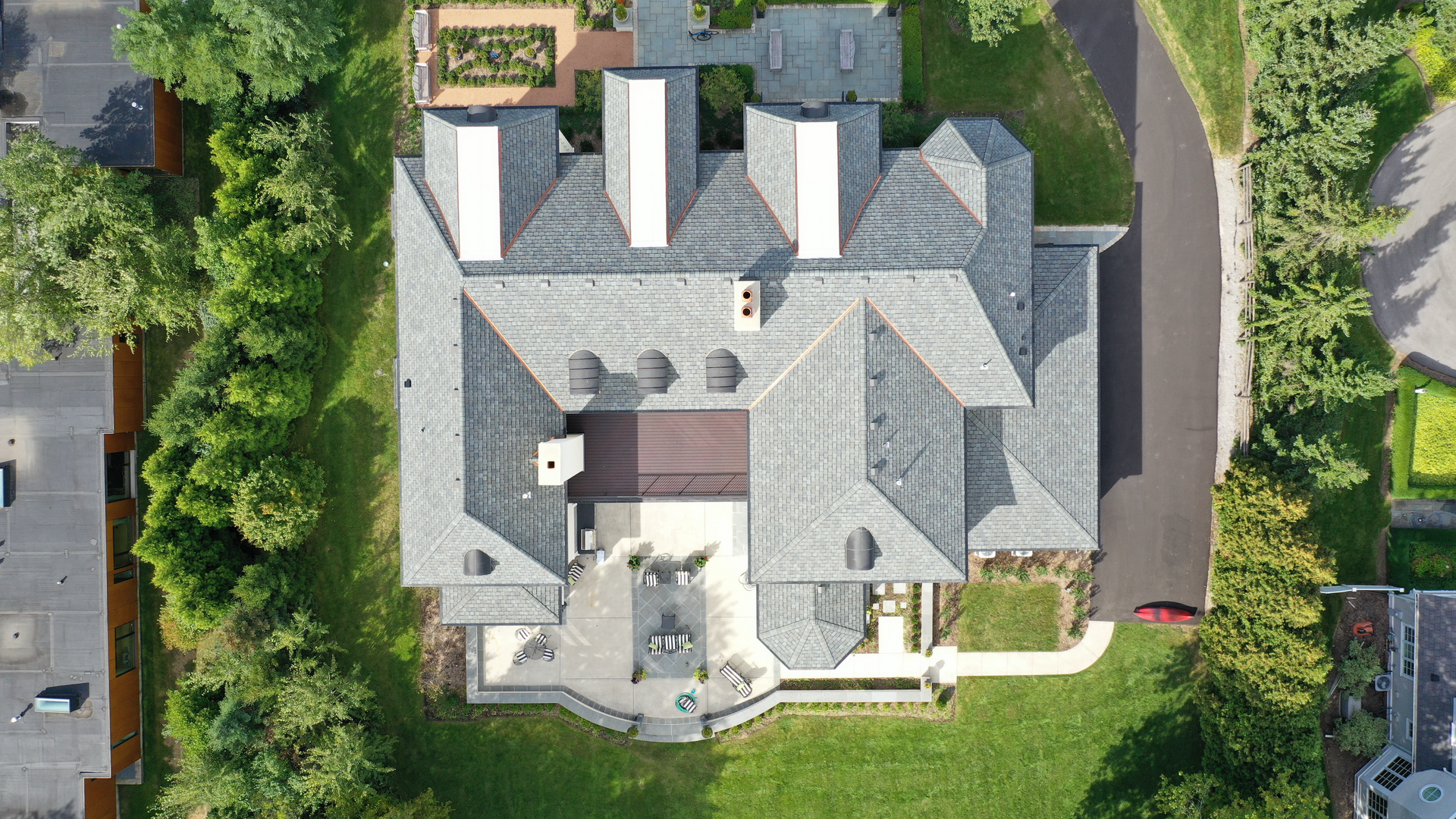 Aerial view of a large house with a gray slate roof, surrounded by a grassy lawn, trees, and a paved driveway.