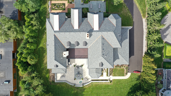 Aerial view of a large house with a gray slate roof, surrounded by a grassy lawn, trees, and a paved driveway.