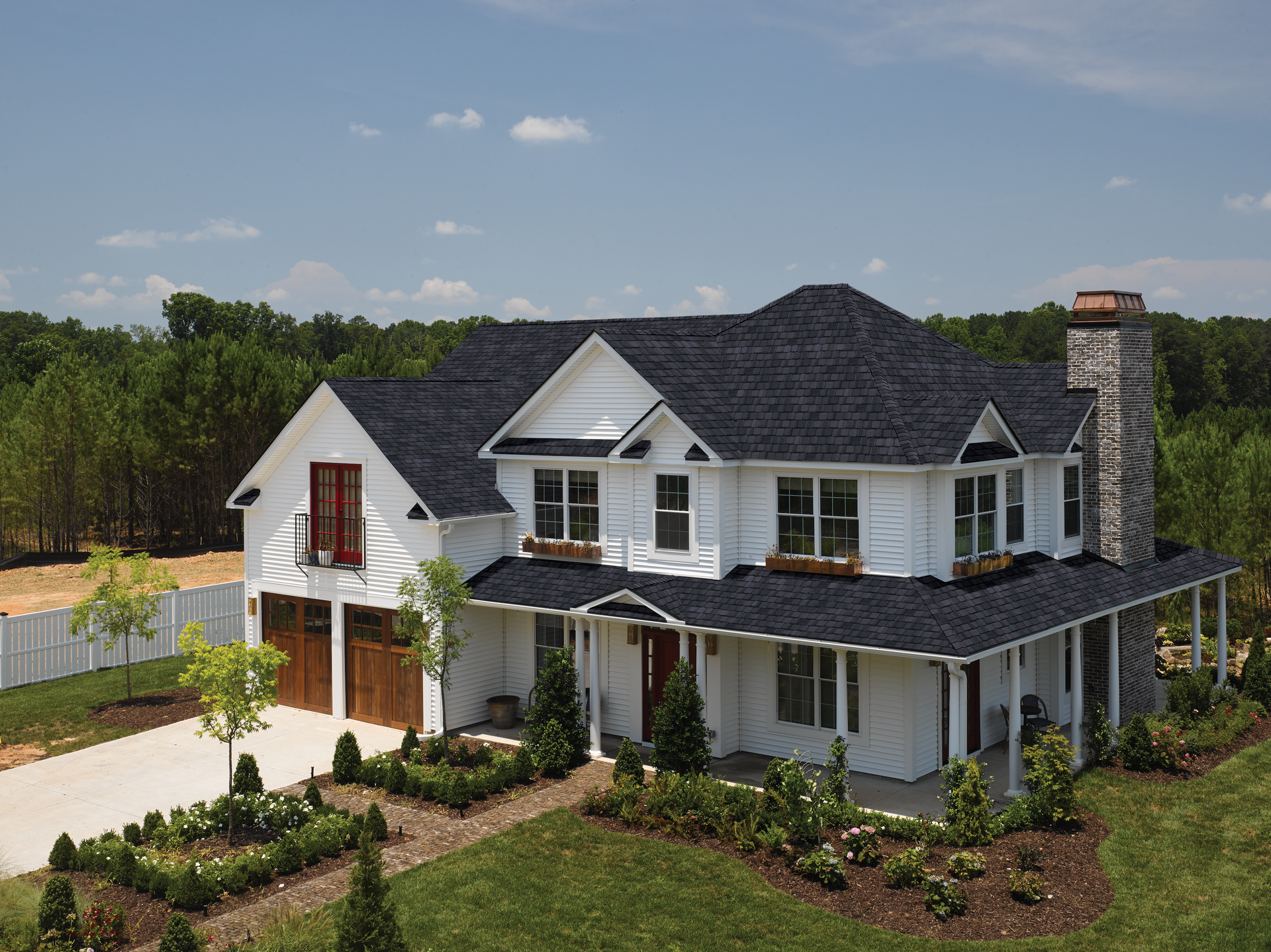 A two-story white farmhouse with a dark shingled roof, stone chimney, and front porch, set in a sunny, green landscape.