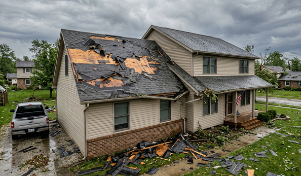 A residential house with significant storm damage, including a partially stripped roof and debris scattered on the lawn.