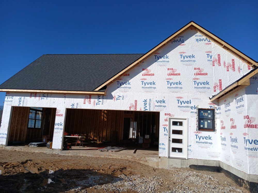 A residential house under construction, covered in white Tyvek wrap, with an open garage and a dark shingled roof.