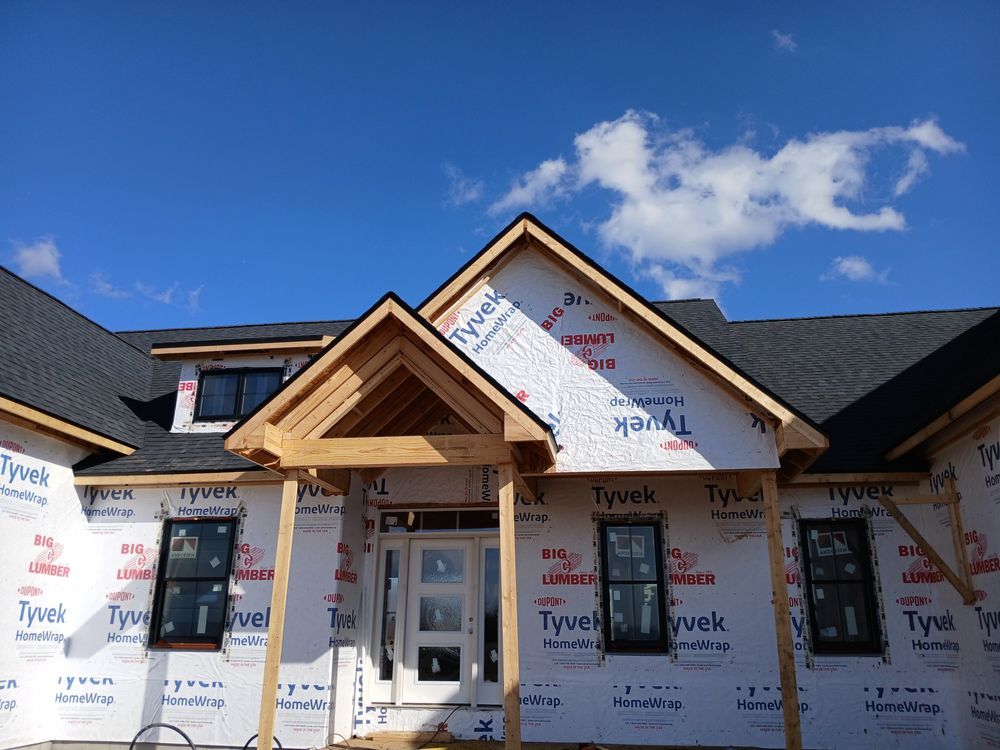 A house under construction with a wooden entrance frame, black-framed windows, and walls wrapped in white Tyvek material.