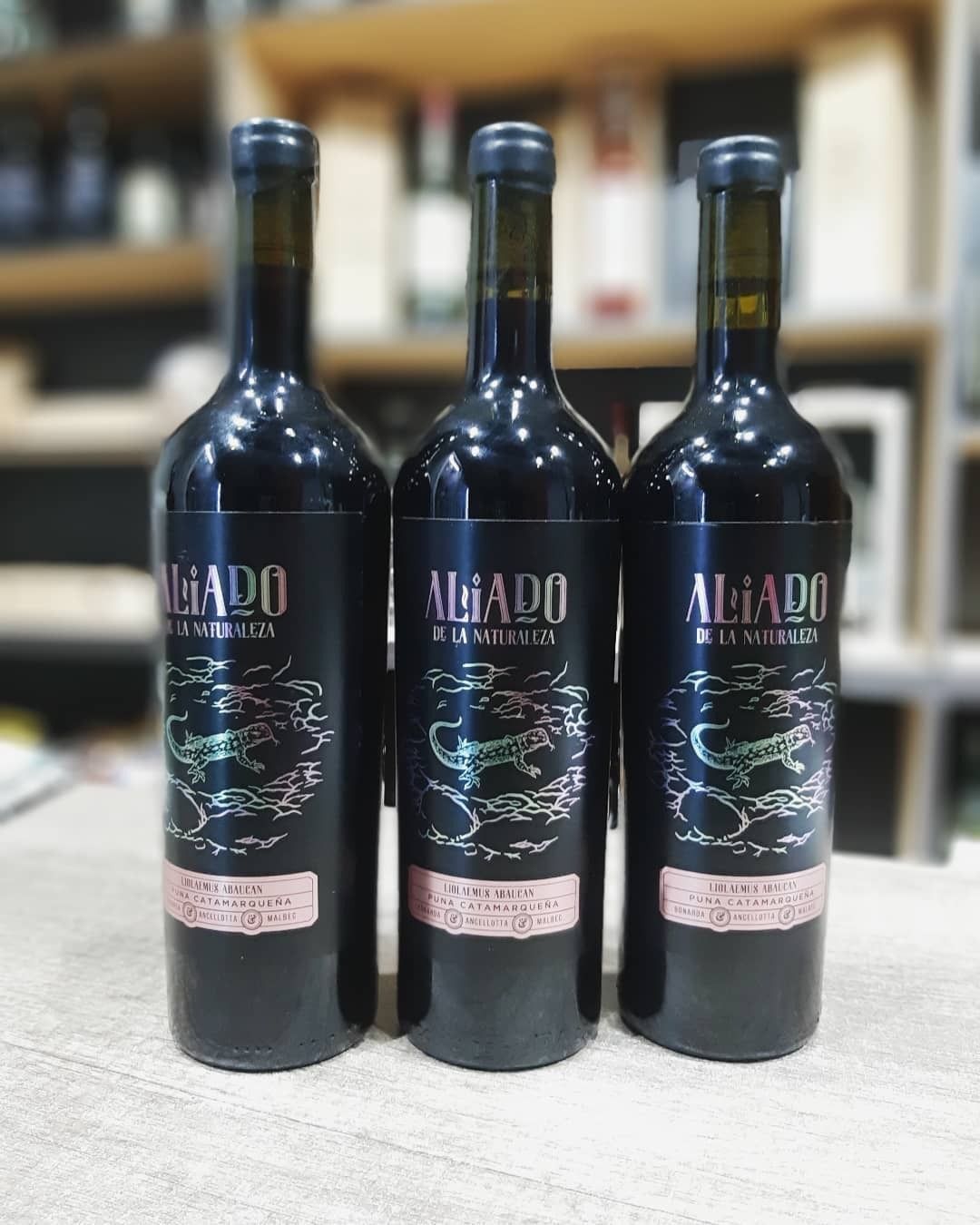 Tres botellas de vino oscuro con etiquetas coloridas sobre una mesa frente a un estante.