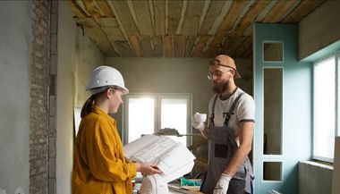A man and a woman are standing in a room looking at blueprints.
