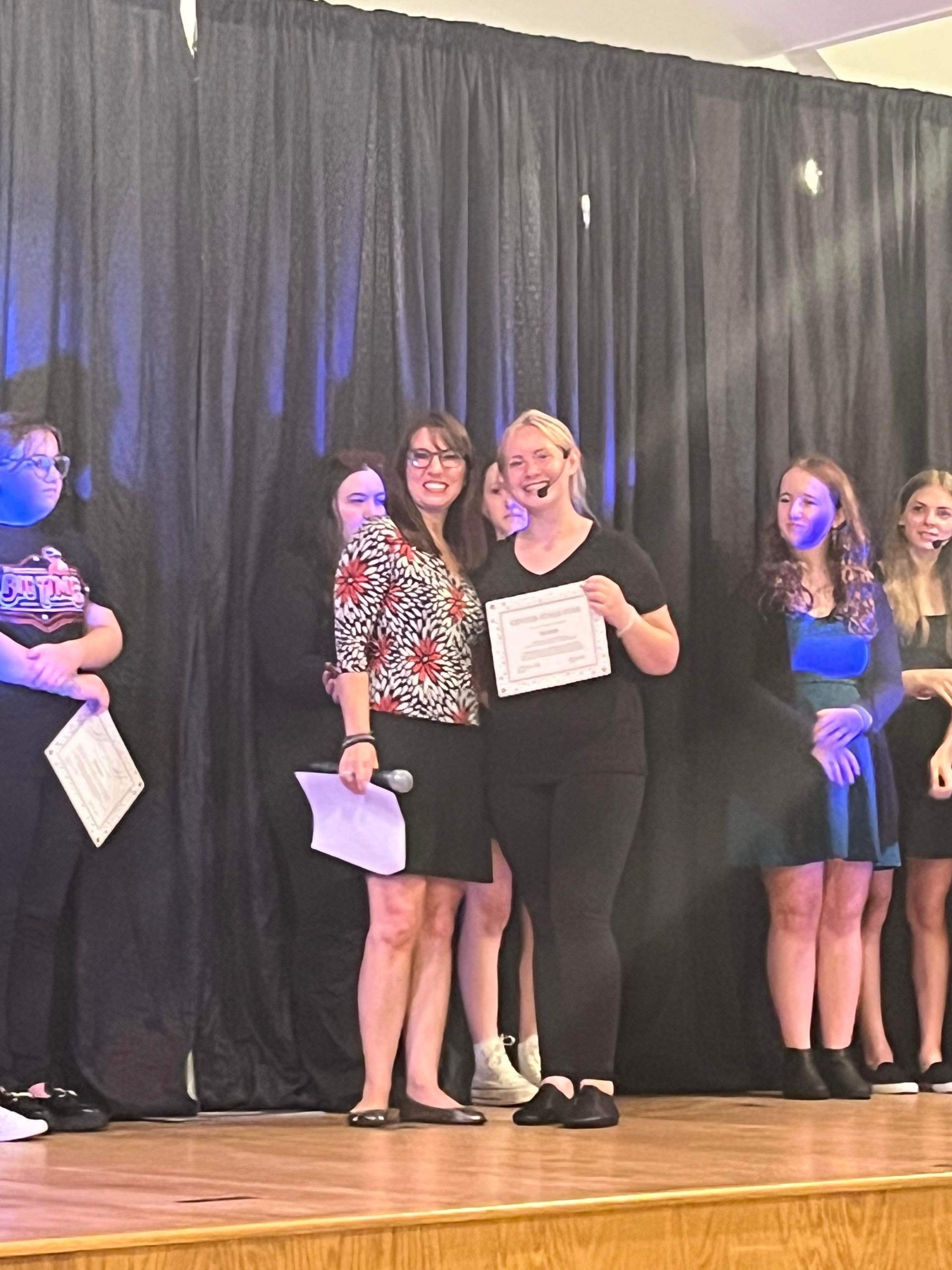 A group of women are standing on a stage holding papers.