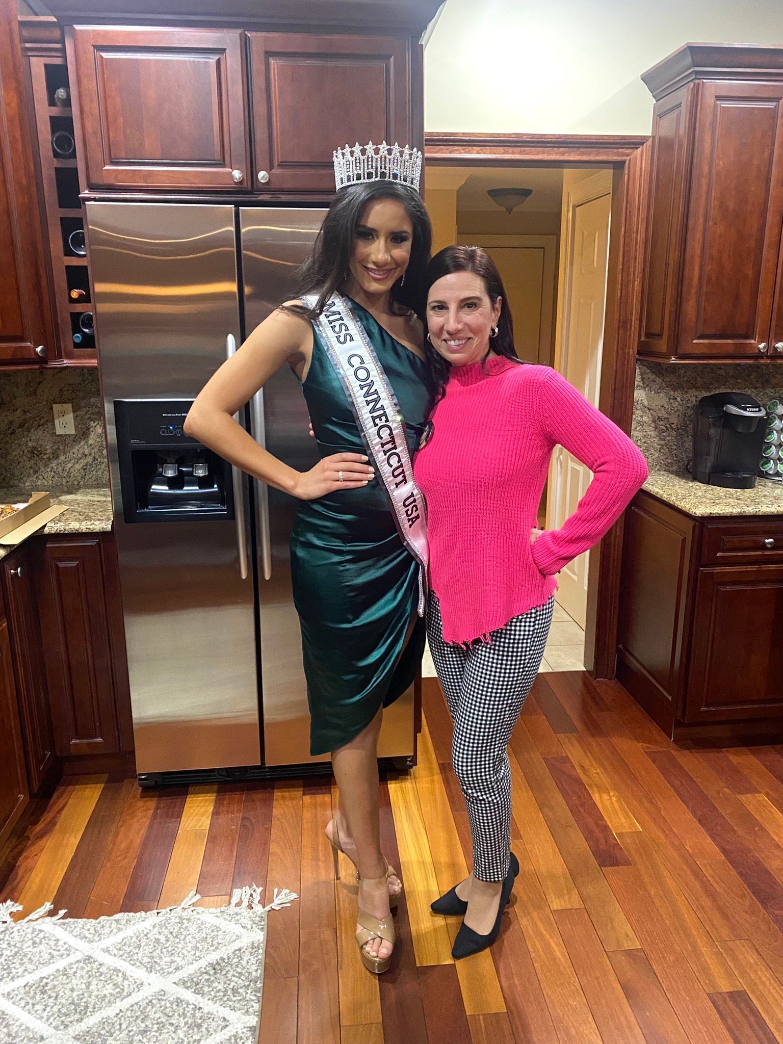 A woman in a green dress is standing next to a woman in a pink sweater in a kitchen.