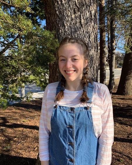 A young woman in overalls is smiling in front of a tree.