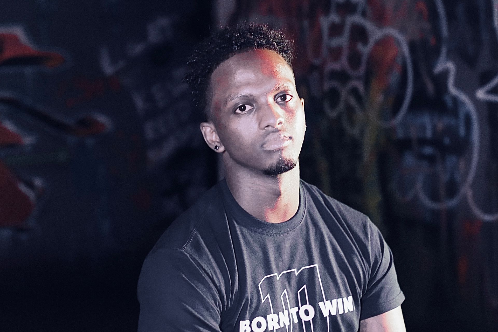 A man wearing a black shirt that says born to win is standing in front of a graffiti wall.