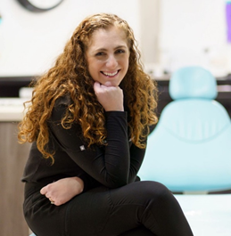 A woman with curly hair is sitting in a dental chair and smiling.