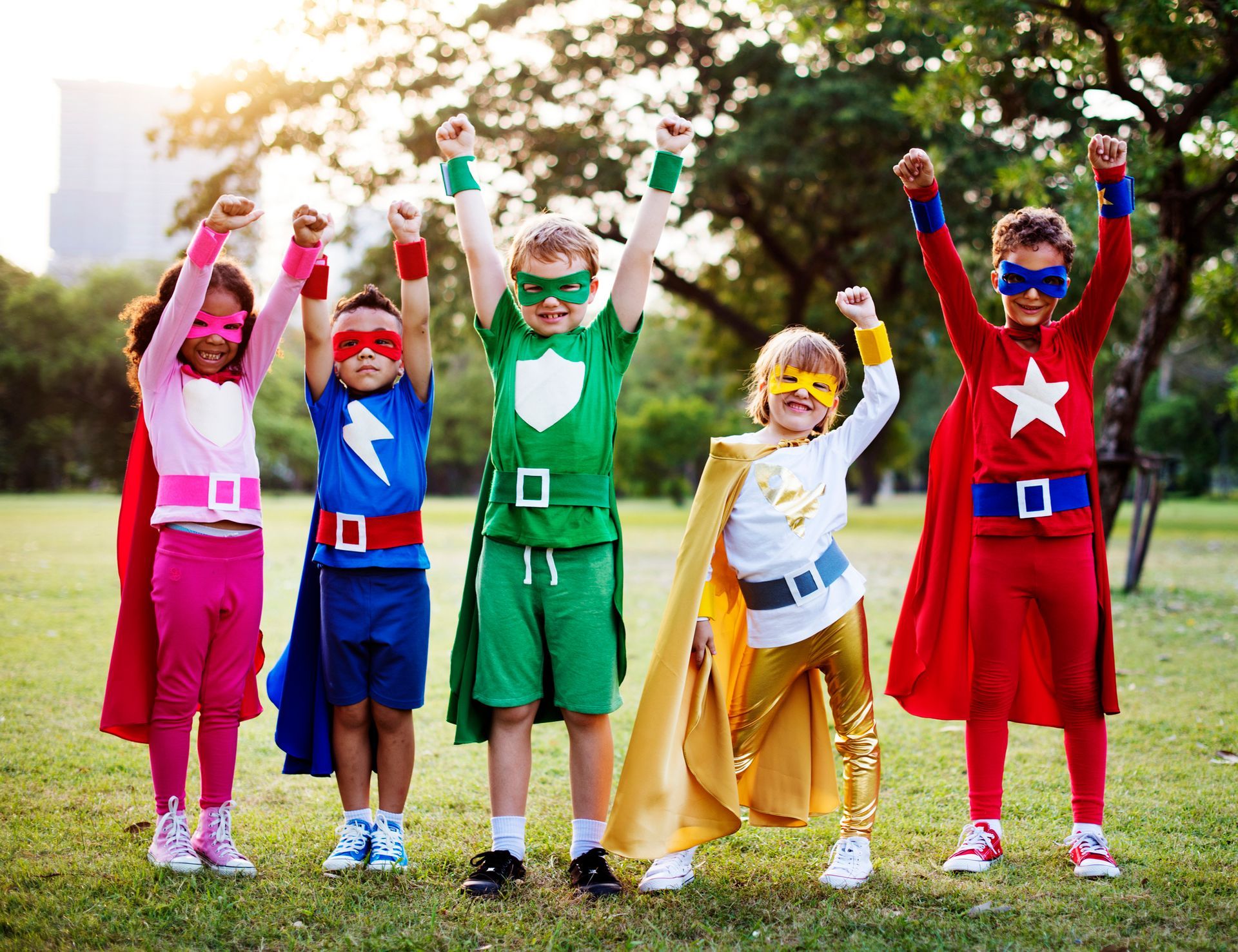 A group of children dressed in superhero costumes are standing in a park.