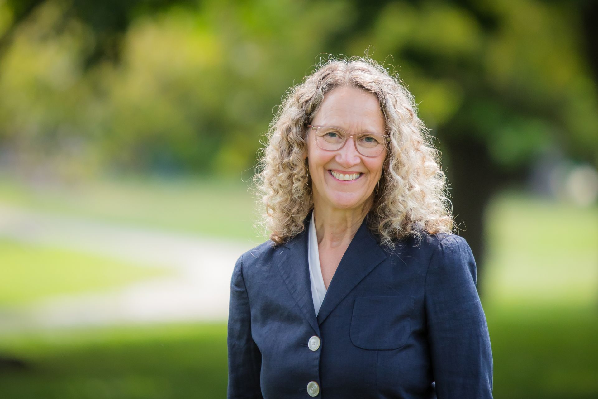 A woman in a blue jacket and glasses is smiling in a park.
