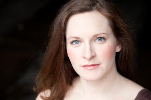 A woman with long red hair and blue eyes is looking at the camera.