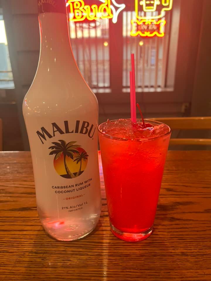 A bottle of malibu rum next to a glass of red liquid on a wooden table.