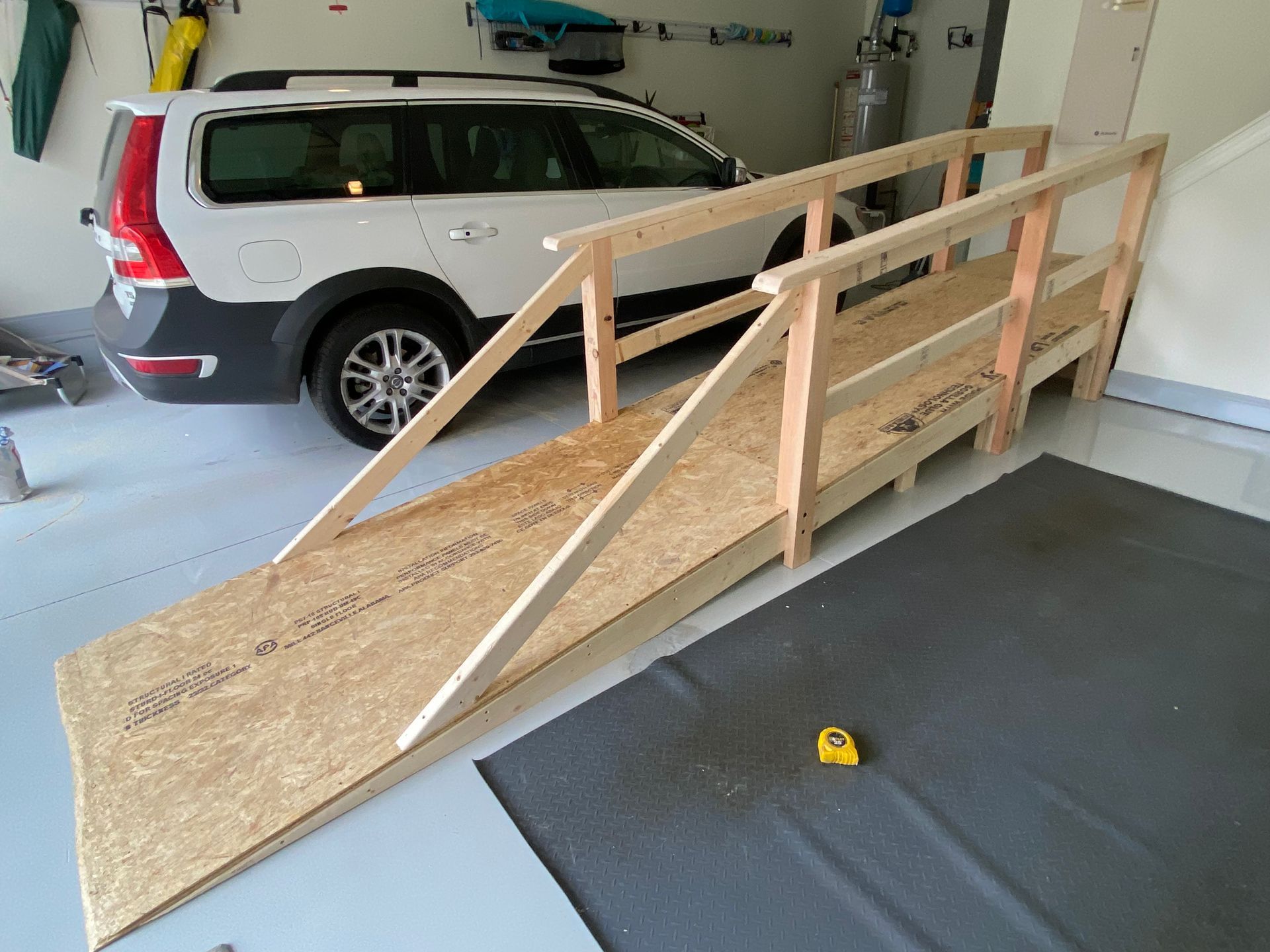 Wooden ramp and deck attached to brick house in a sunny backyard; person walking at left.