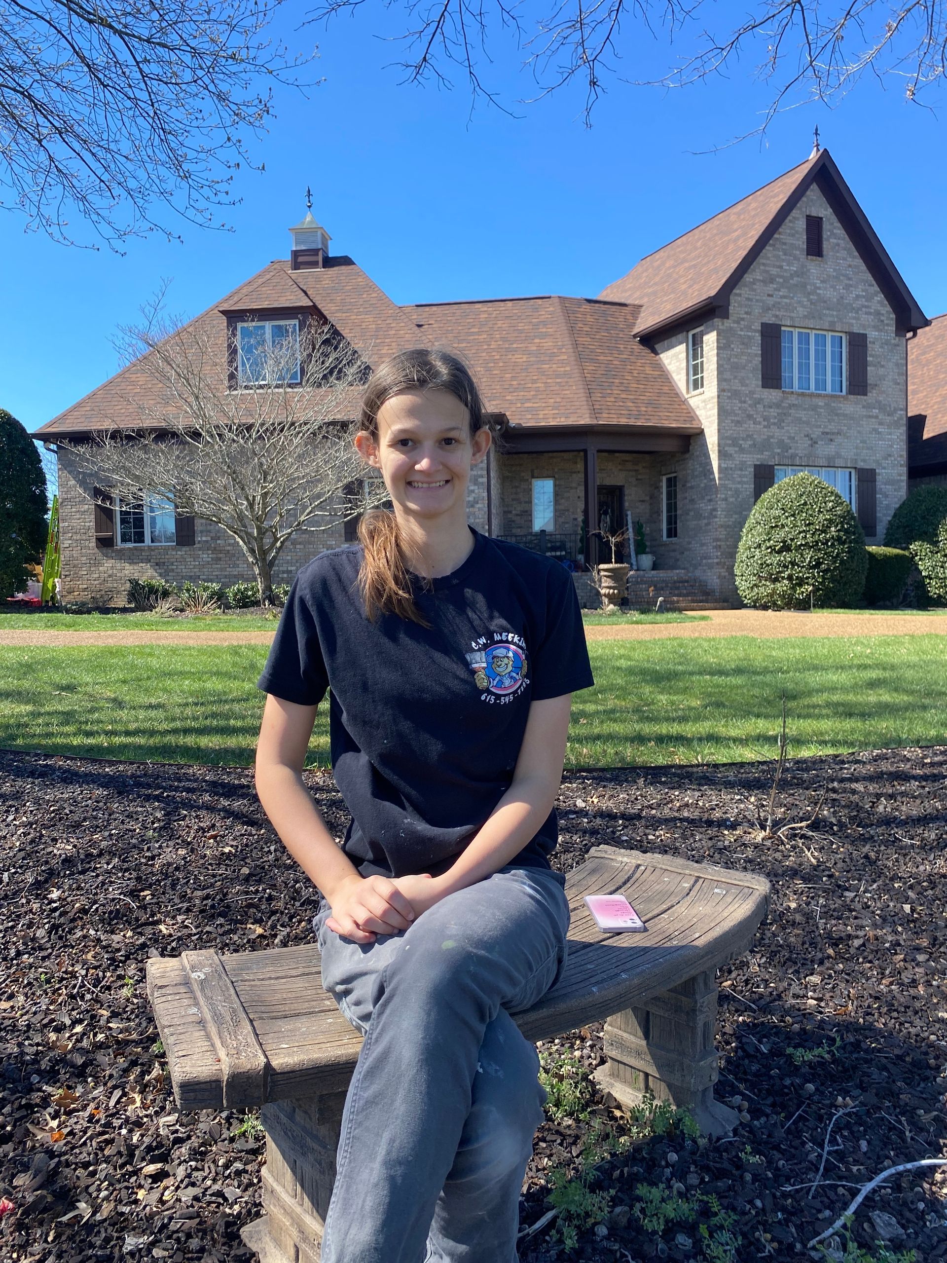 A person sits on a stone bench outdoors, smiling, with a large, light-colored brick house in the background.