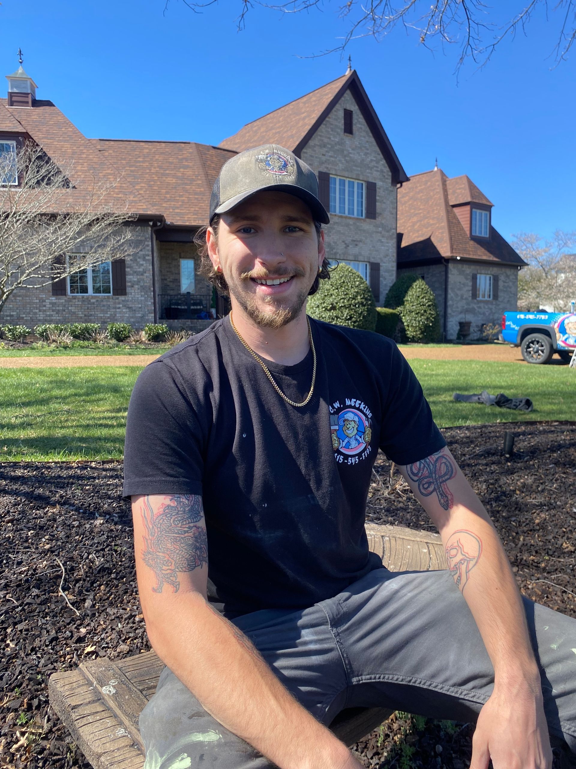A person wearing a baseball cap and a dark t-shirt sits on the ground in front of a large brick house on a sunny day.