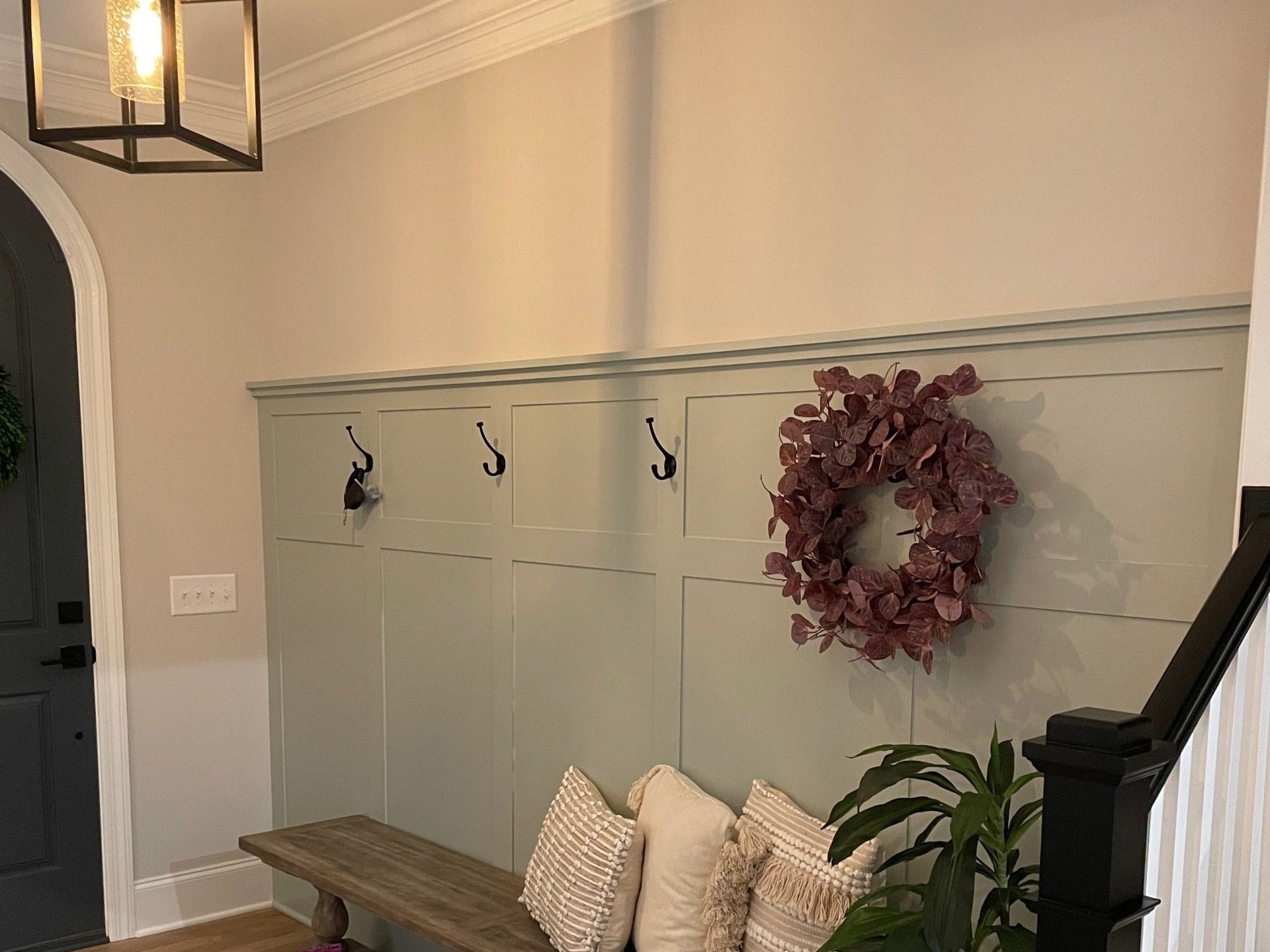 Entryway with pale green wainscoting, wall hooks, a wreath, bench with pillows, and a black lantern light fixture