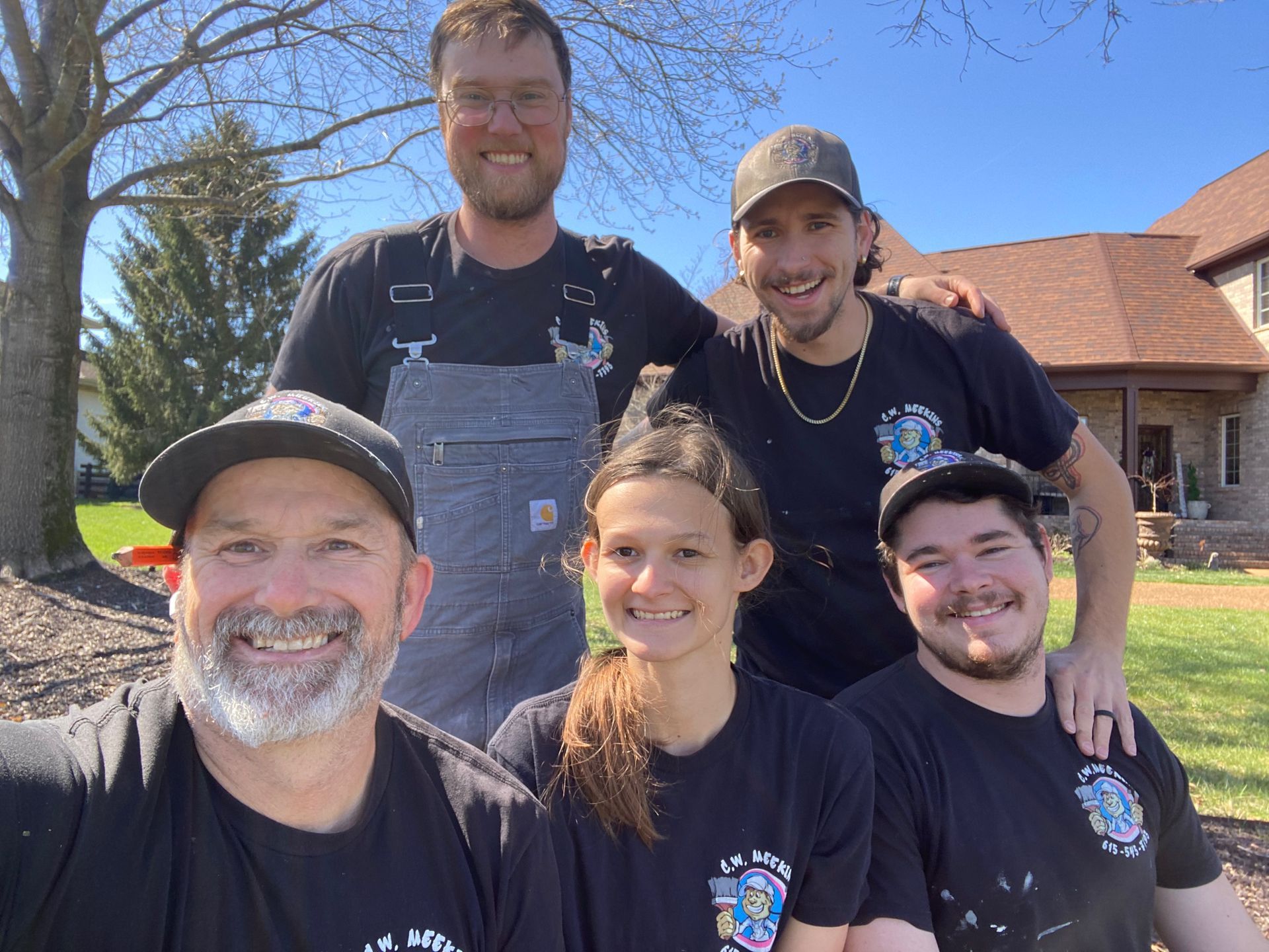 A group of five people wearing black shirts pose for a smiling selfie outdoors in front of a brick house.