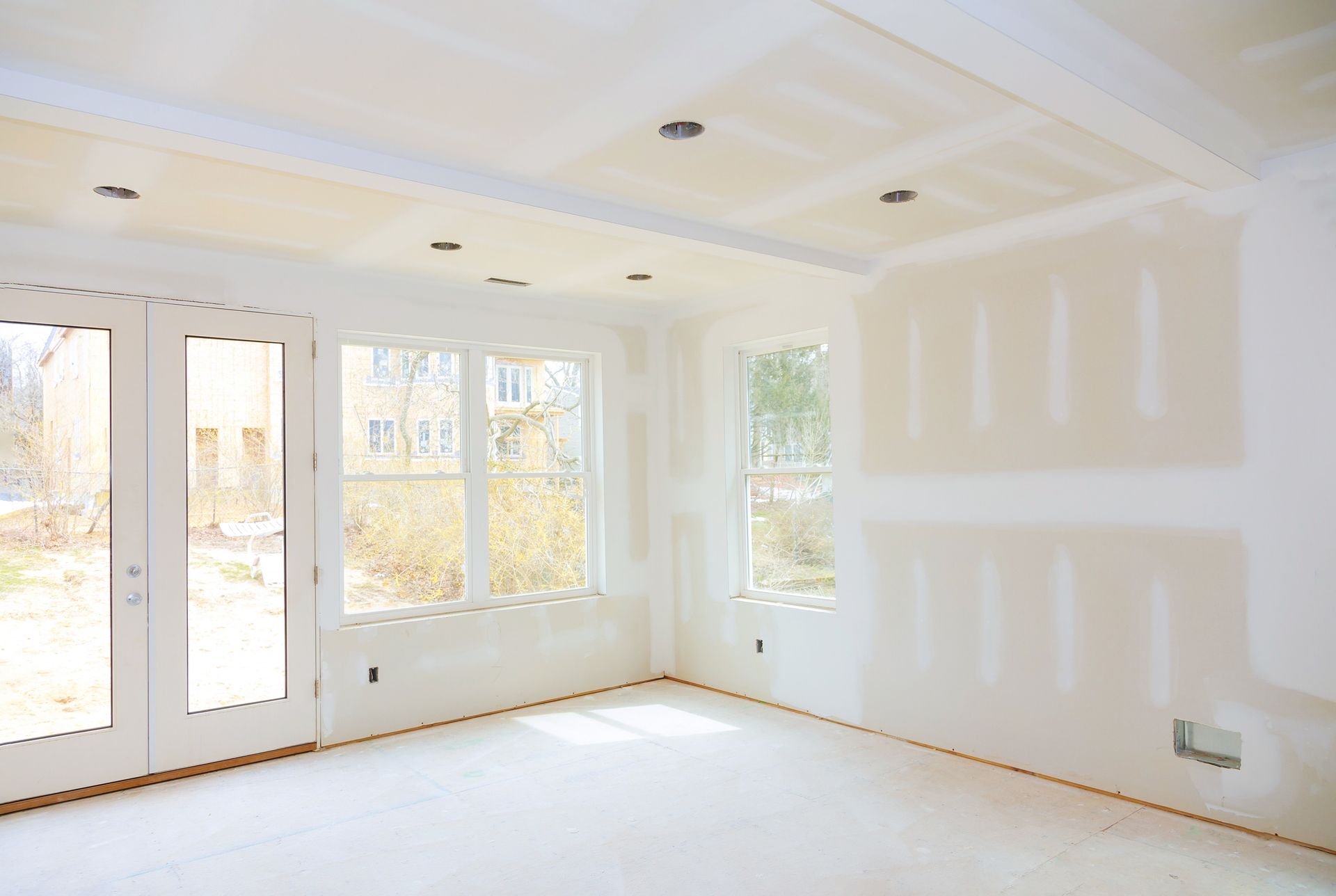 Bright empty room with white walls, large windows, and unfinished drywall patches