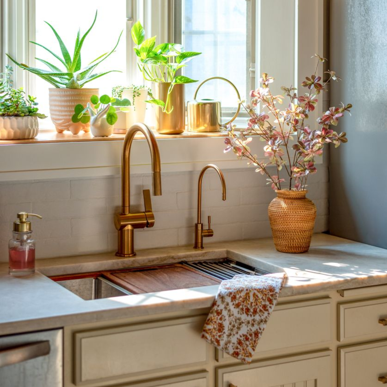 A kitchen sink with gold fixtures, a wooden cutting board, a woven vase with floral stems, and potted plants on the sill.