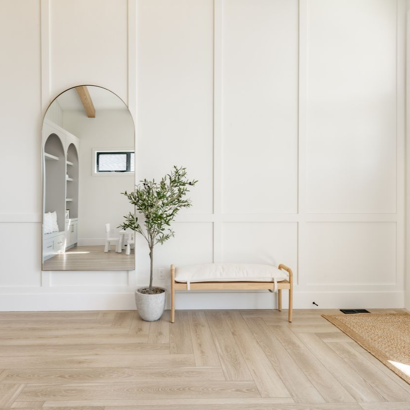 An arched floor mirror, a small potted tree, and a wooden bench with a white cushion against a white paneled wall.