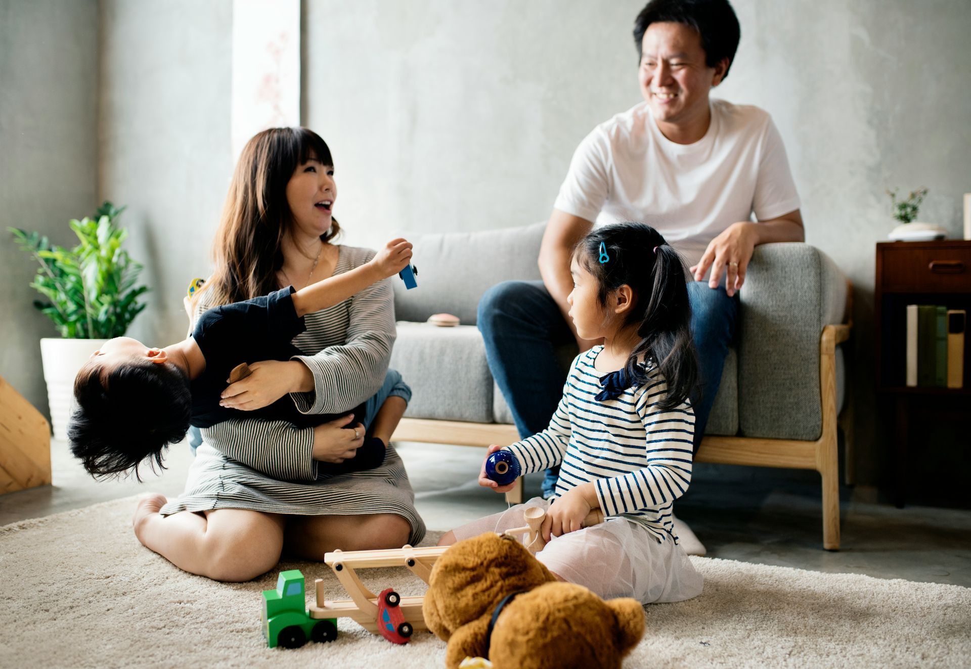 Family playing in a living room: mother holding child, father on couch, child playing with toys.