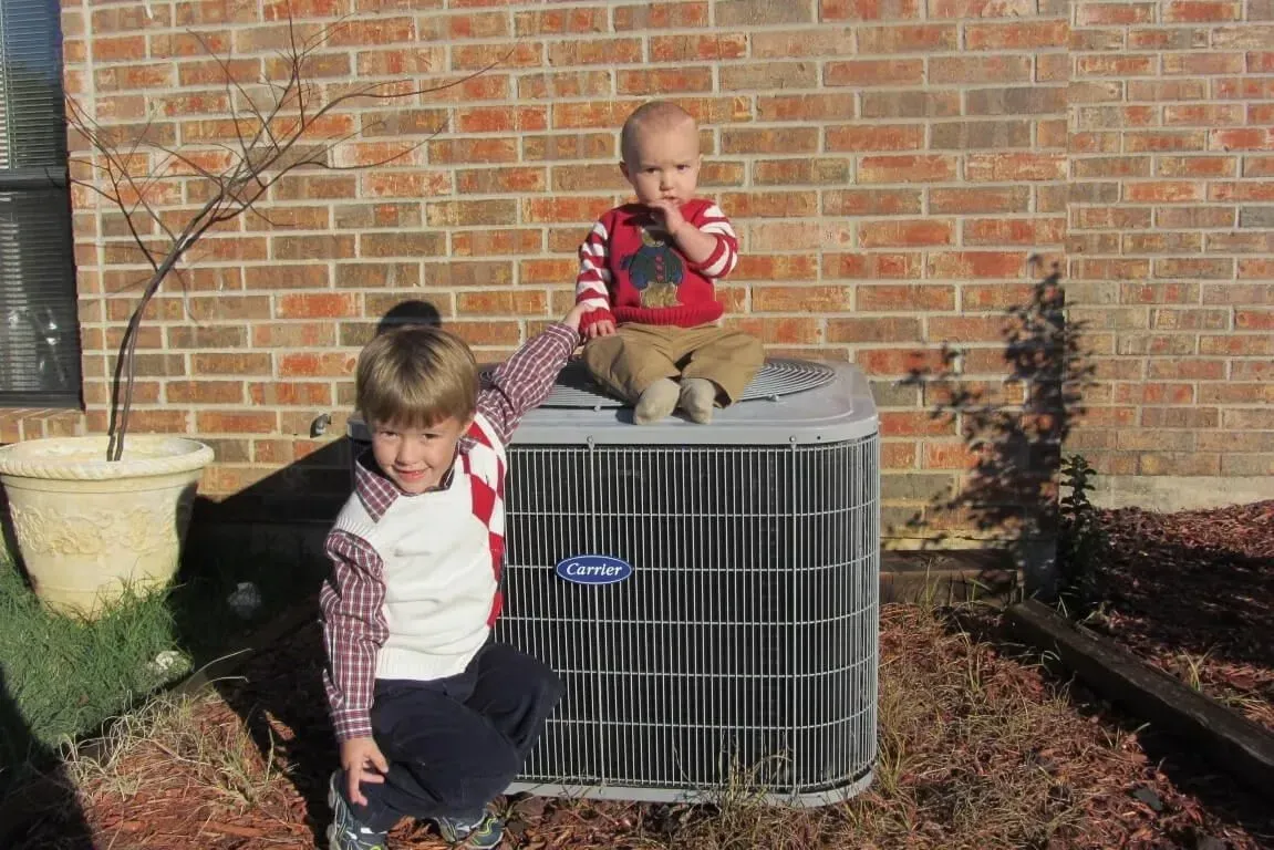 Two children on an air conditioner unit in front of a brick wall. One child points, the other looks on.