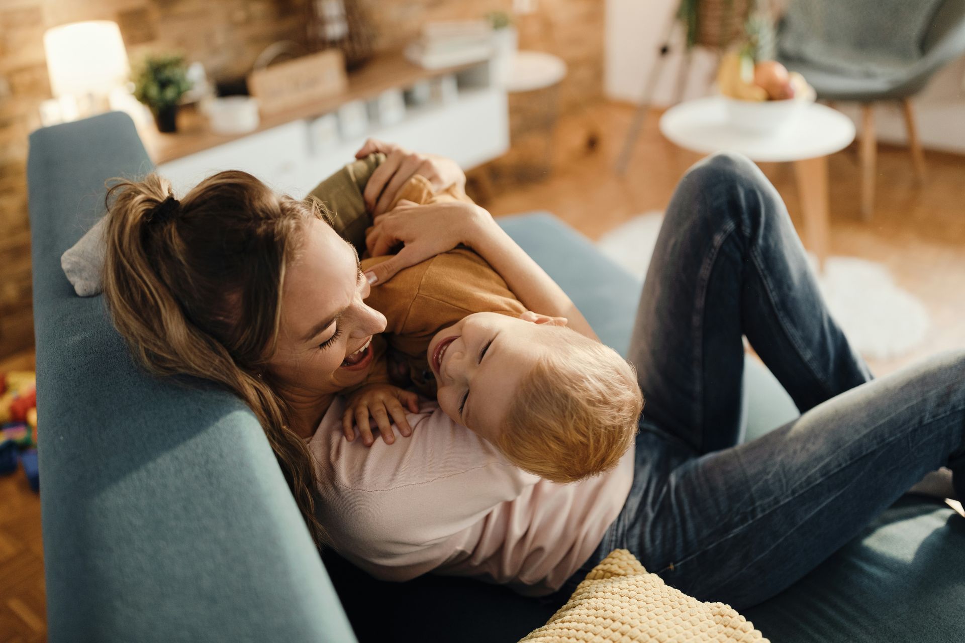 Woman and child laughing, playing on a blue couch in a living room.
