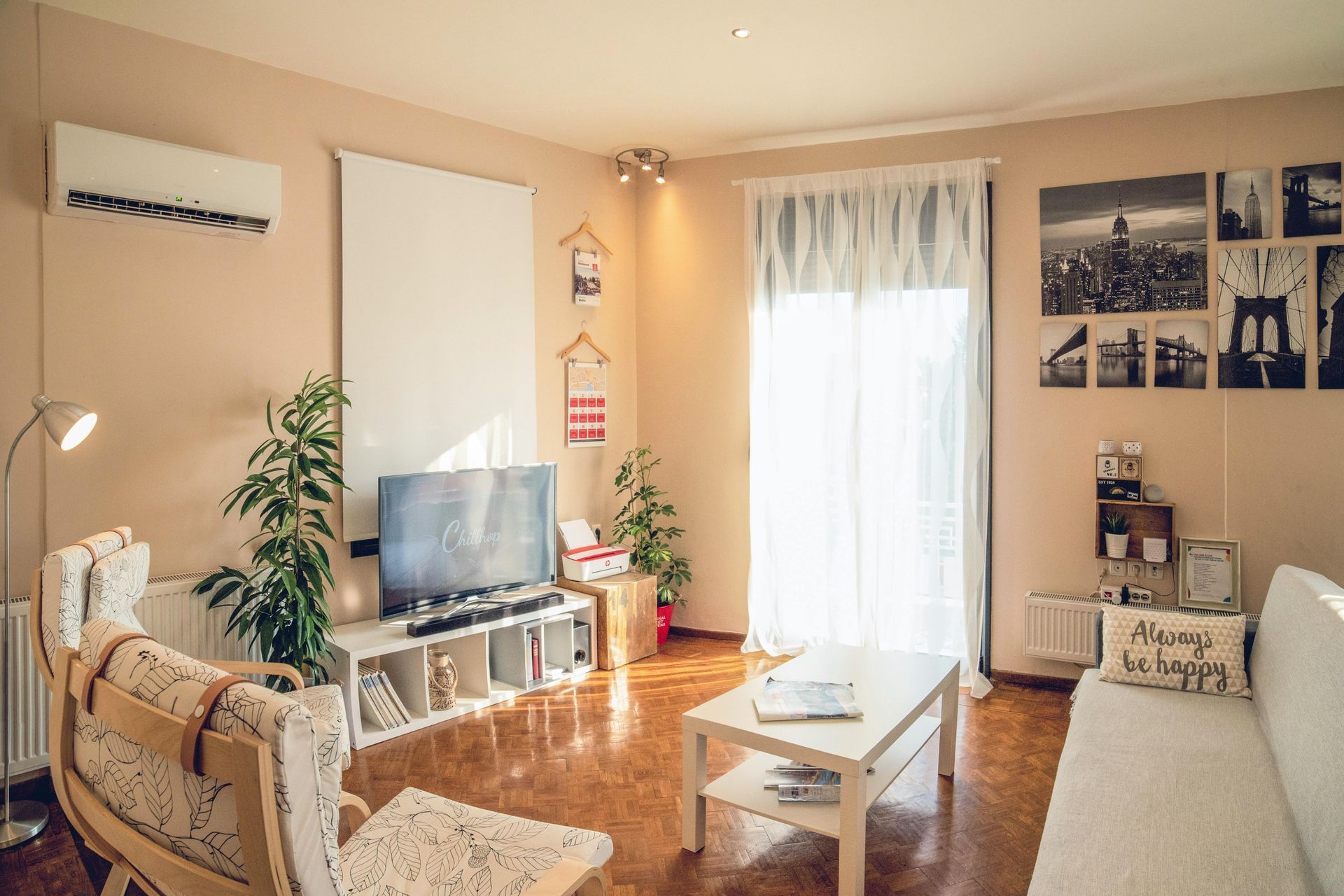 Living room with white furniture, plants, TV, and sunlight streaming through a sheer curtain.