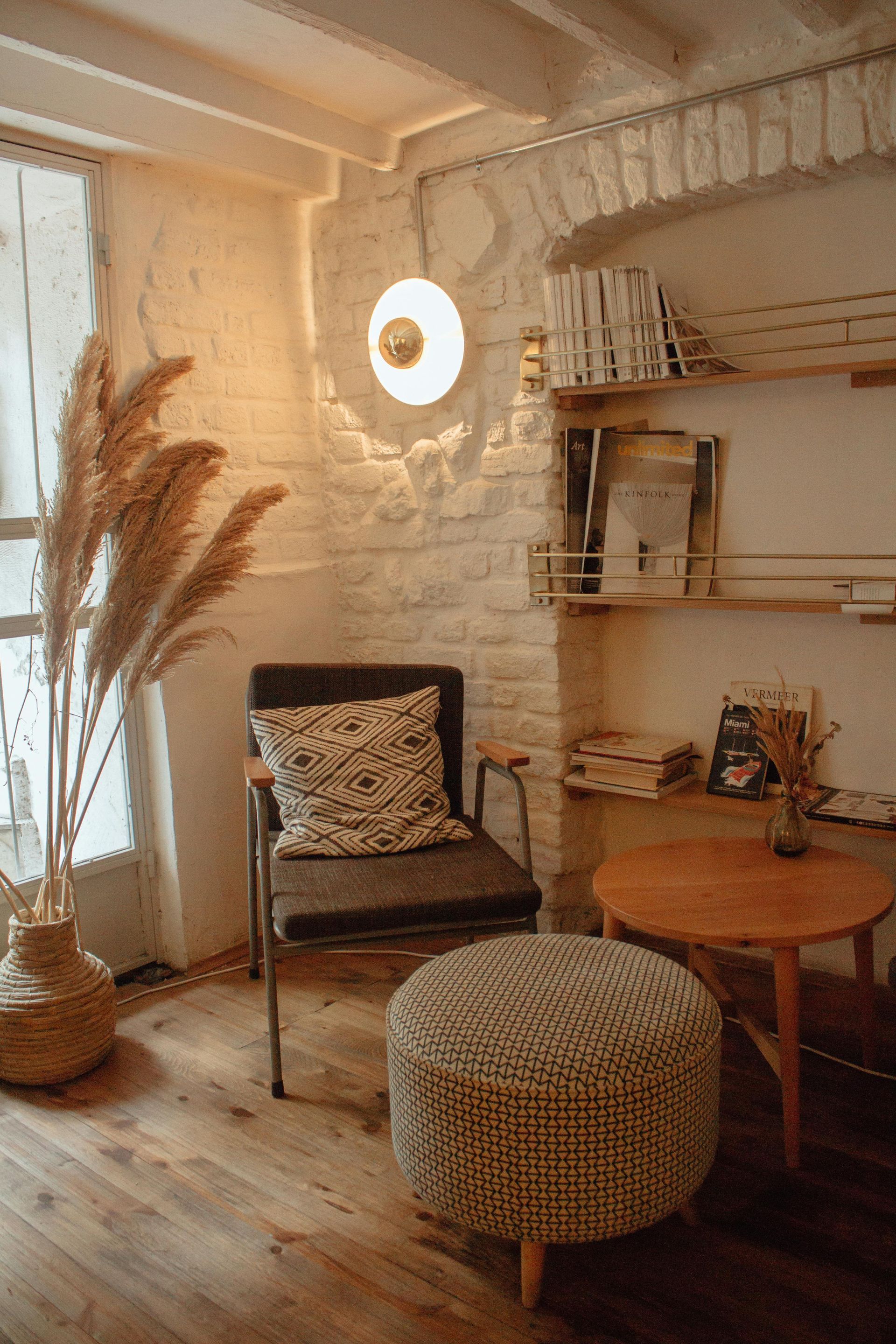 Cozy corner with a chair, round ottoman, and small wooden table. Dried pampas grass in a vase near a window.