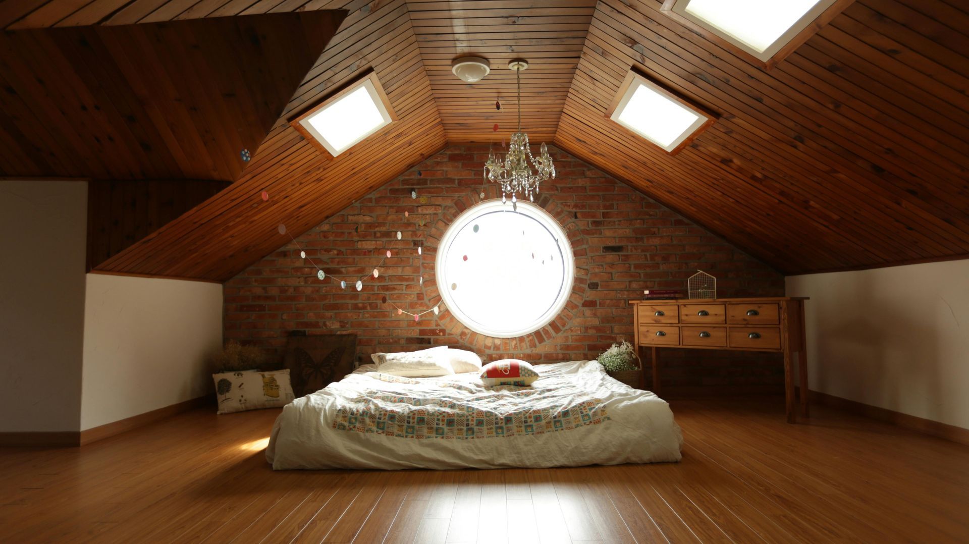 Cozy attic bedroom with bed, round window, wooden ceiling, brick wall, and skylights.
