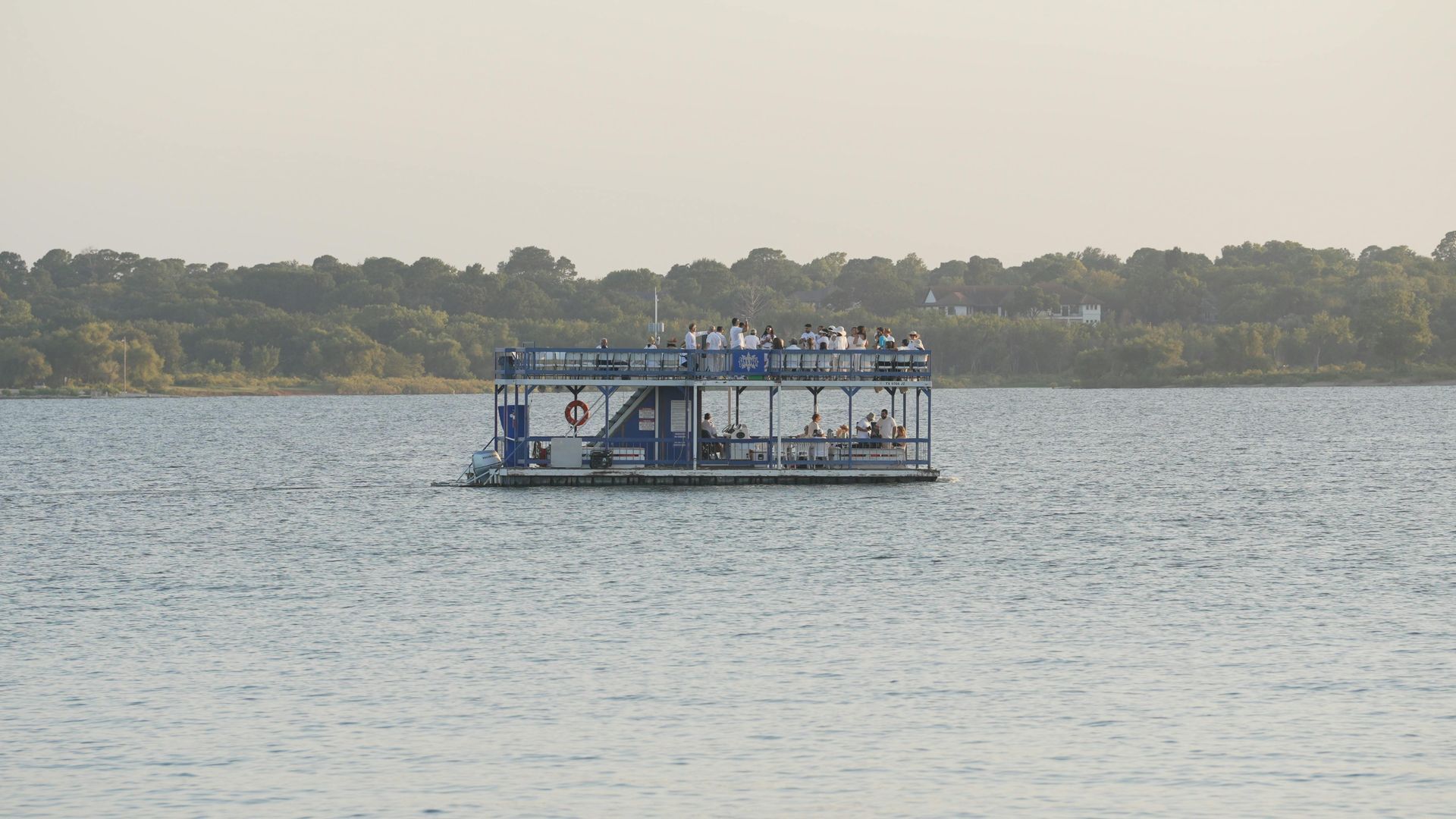 Boat on water with people on board; blue and white color scheme; trees in background.