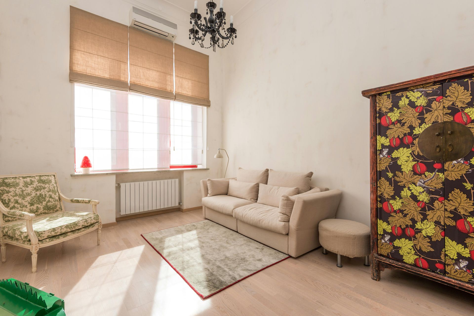 Living room with sofa, chair, and patterned cabinet; window with shade, chandelier, and rug.
