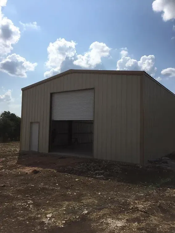 A large metal building with a garage door is sitting in the middle of a dirt field.