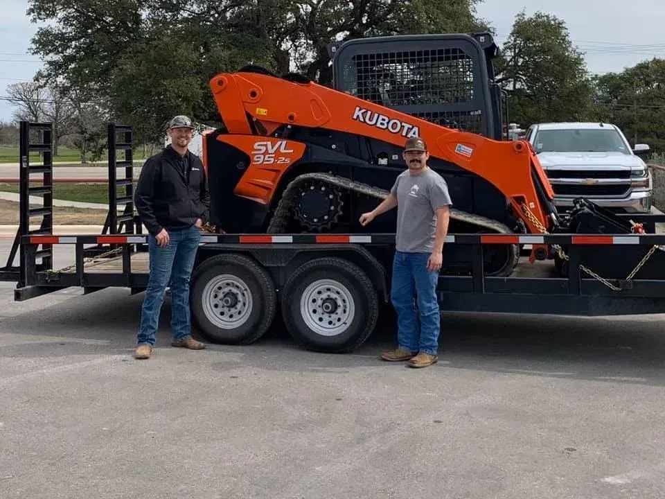 Two men are standing next to a trailer with a kubota skid steer on it.