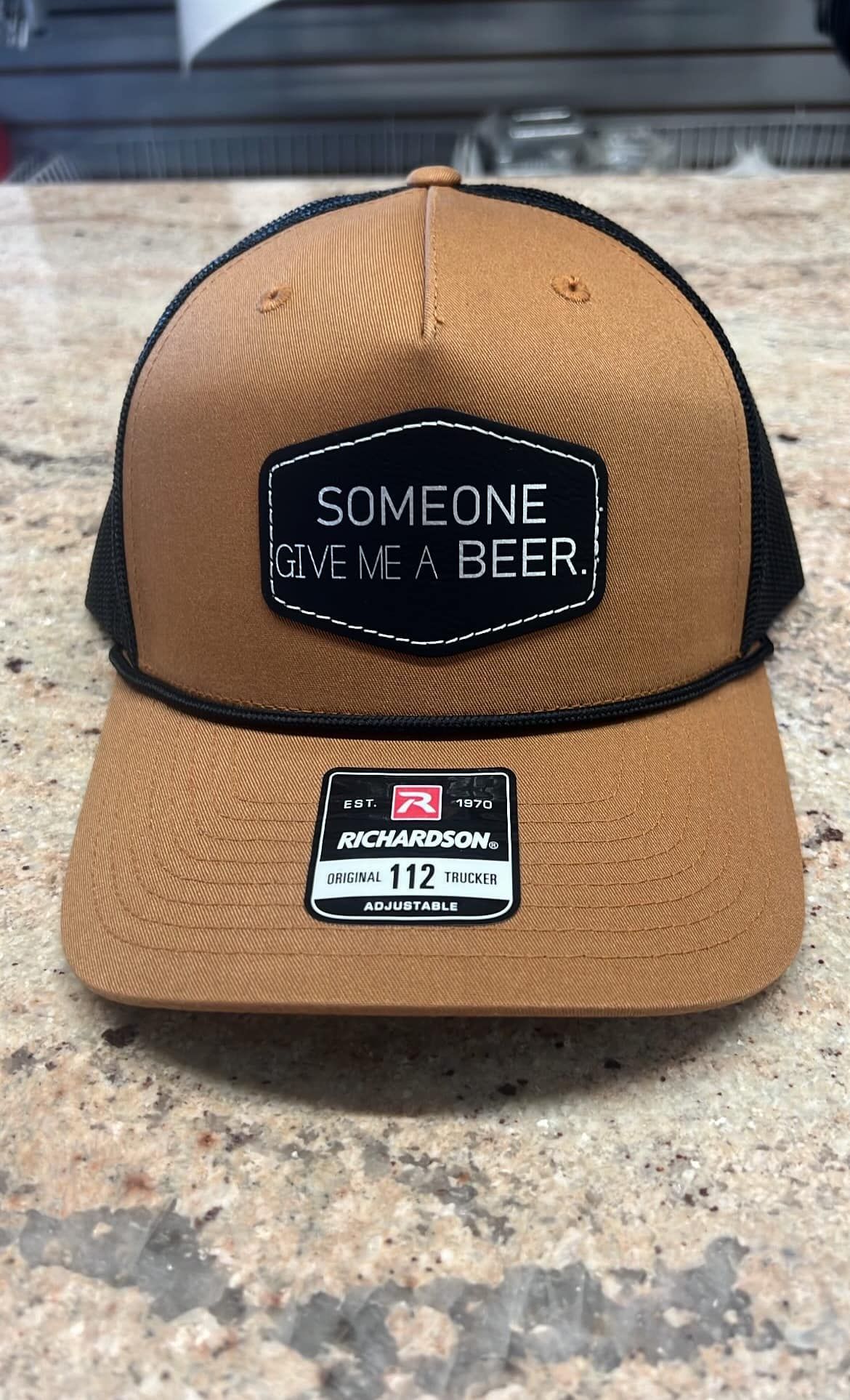 A baseball cap with a patch that says `` someone give me a beer '' is sitting on a counter.