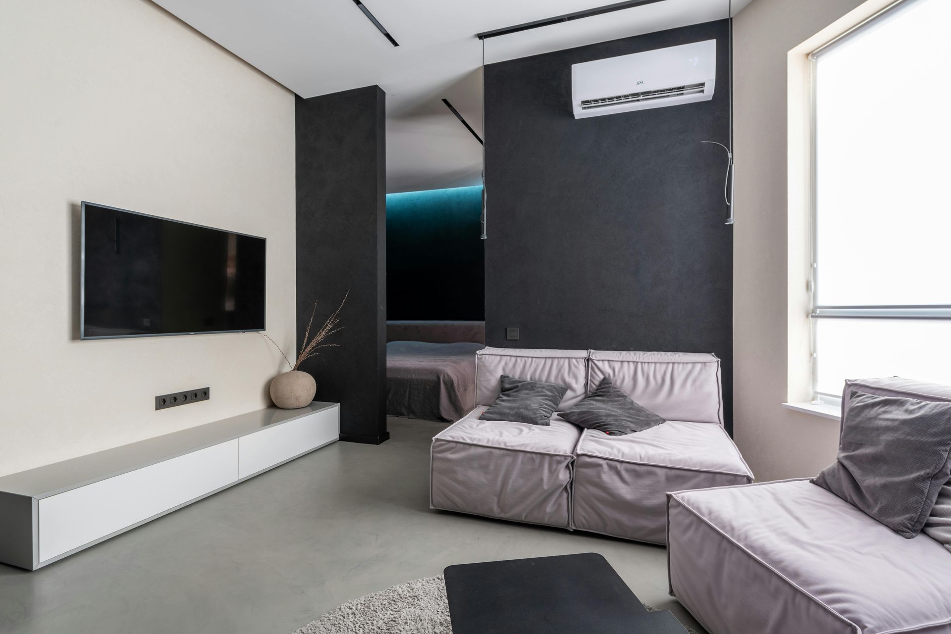 Modern living room with light gray couch, TV, and dark accent wall. There's a window, white cabinetry, and a view of a bed in the background.