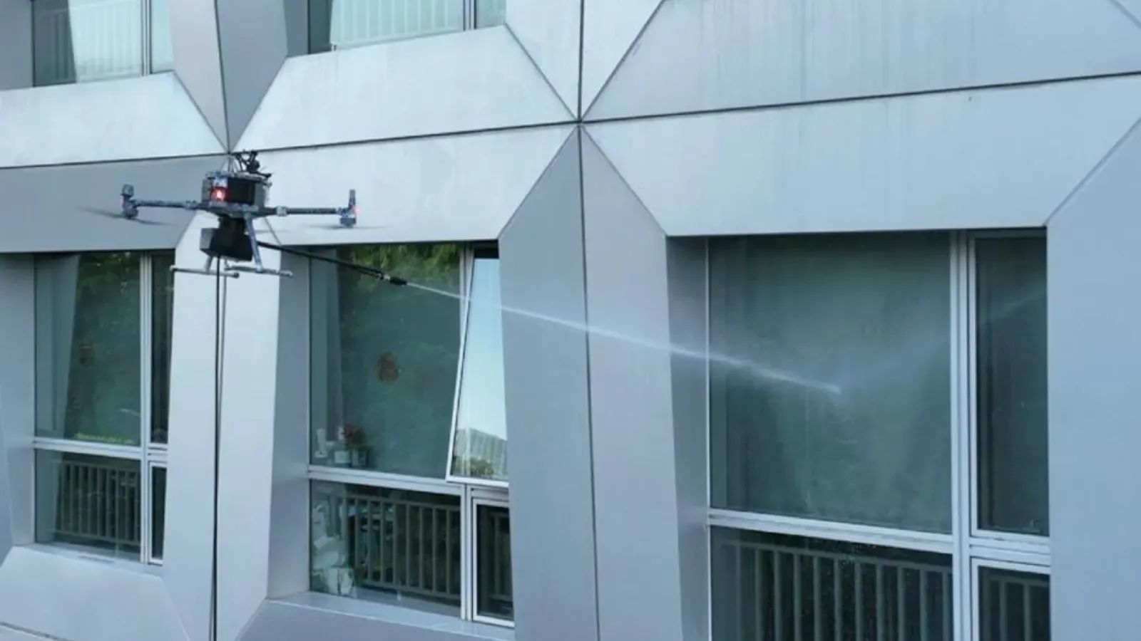 Drone spraying a building facade with water; gray building with geometric design, sunny day.