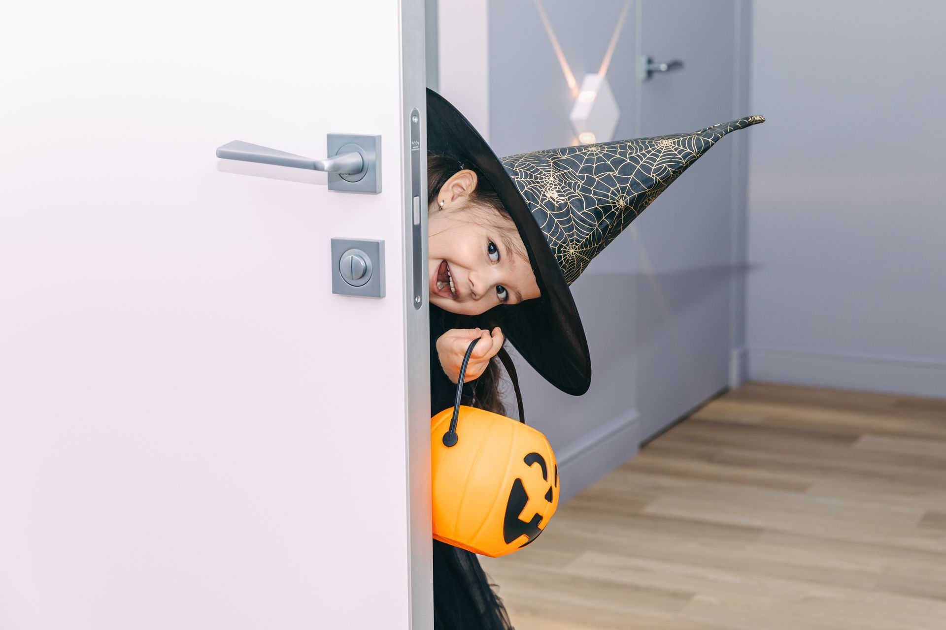 Child dressed as a witch peeking from a door holding a pumpkin bucket, smiling.