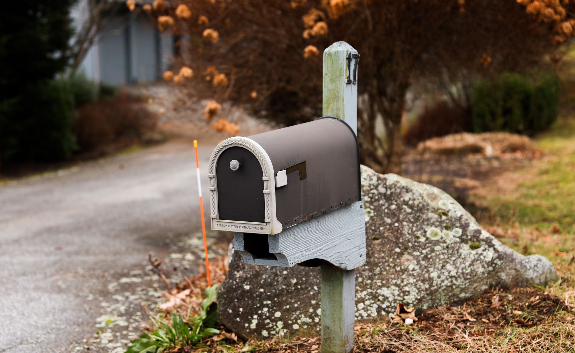 Black mailbox on a concrete post, in front of a house, and a large rock.