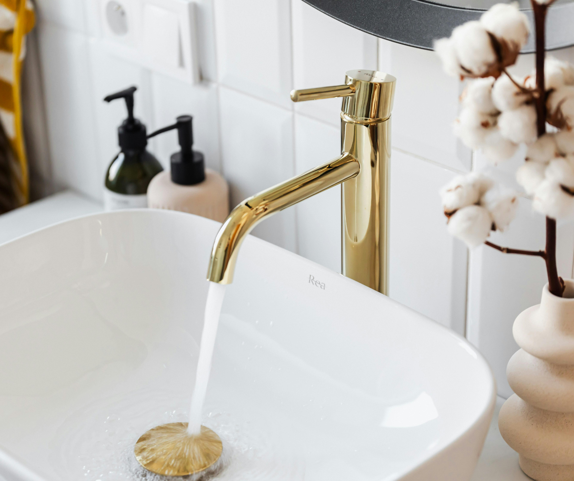 Gold faucet pouring water into a white sink, with soap dispensers and cotton plant in the background.