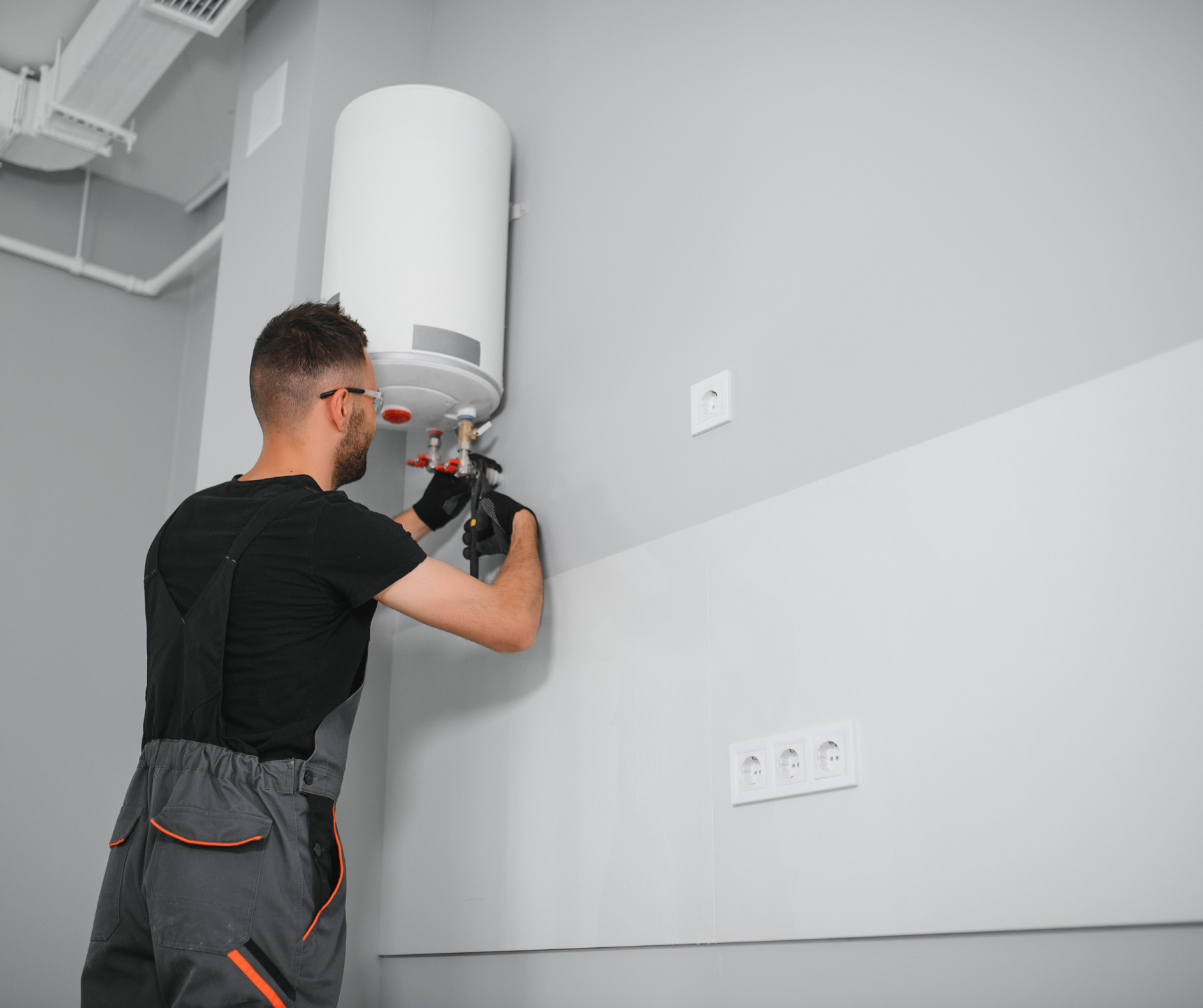 Plumber installing a white water heater on a gray wall, wearing gray overalls and safety glasses.