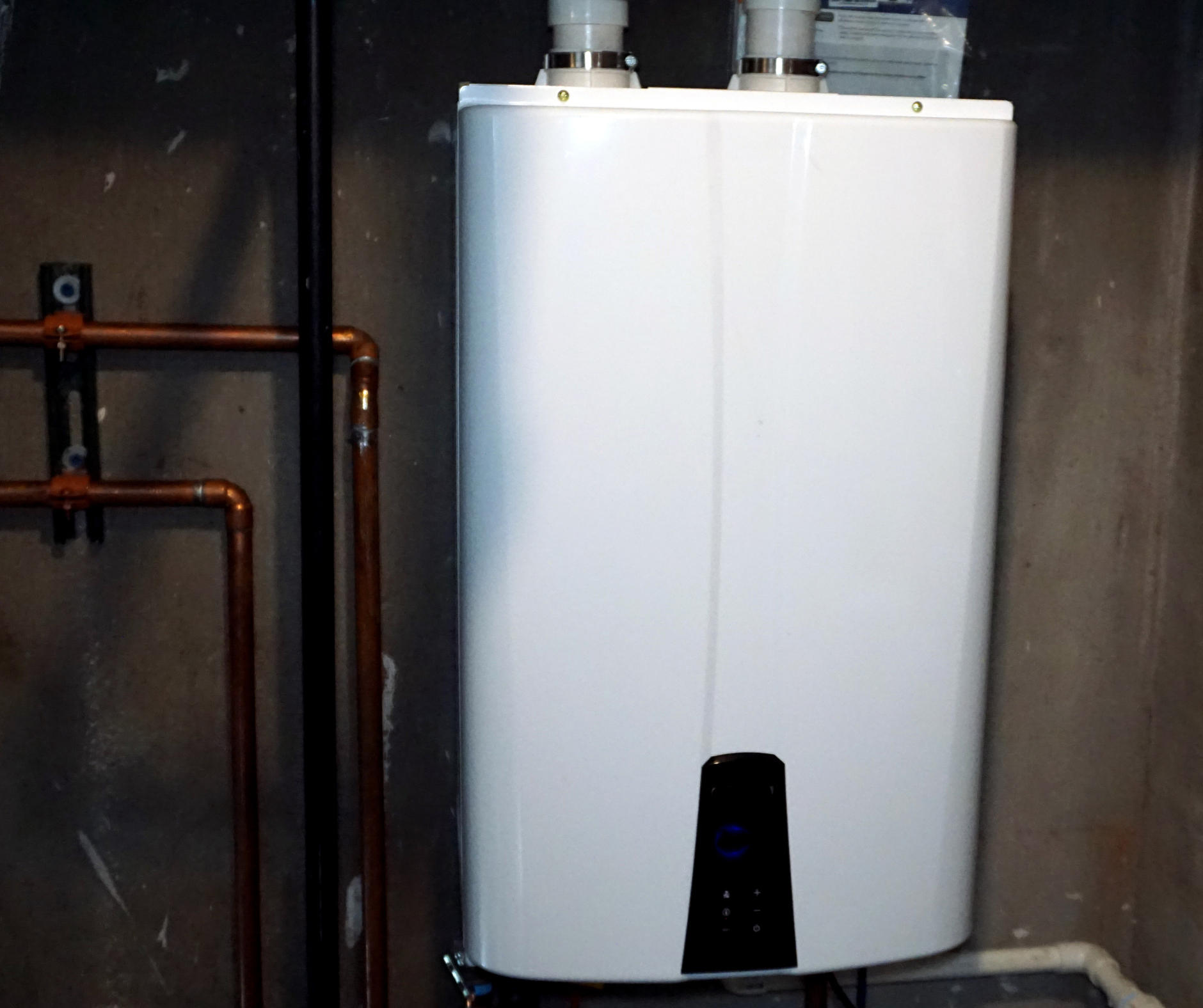 White tank water heater with copper pipes on a dark wall.
