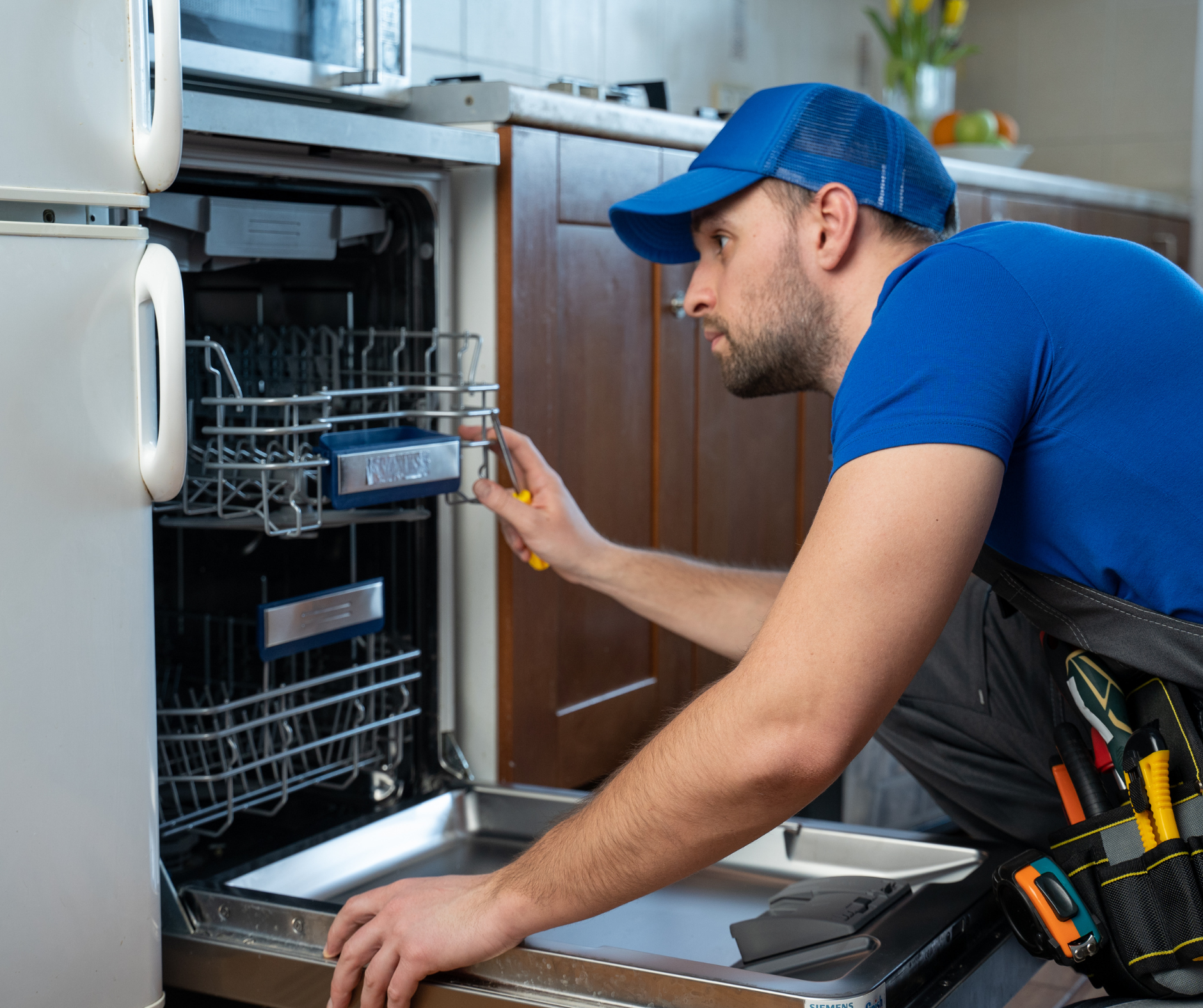 A person in a blue shirt and hat repairs a dishwasher with tools in a kitchen setting.